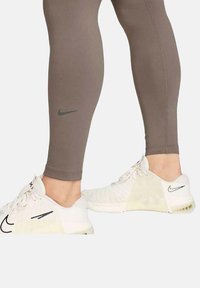 Leggings Nike de couleur marron avec un logo gris, associés à des baskets blanches avec des accents noirs et une tige en maille. Texture lisse, design ajusté.