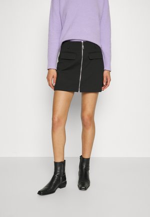 Kobieta ubrana w lawendowy sweter, czarną mini spódnicę na zamek z kieszeniami oraz czarne botki, stoi na tle białego tła.