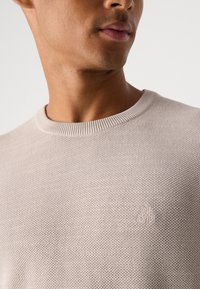 Pull en maille beige clair avec un motif texturé, col rond côtelé, et un logo en relief subtile sur la poitrine. Fini en tissu doux.