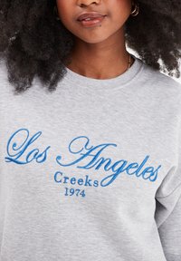 Felpa grigia chiara con ricamo blu che recita "Los Angeles Creeks 1974." Design a collo rotondo con texture morbida e vestibilità rilassata.