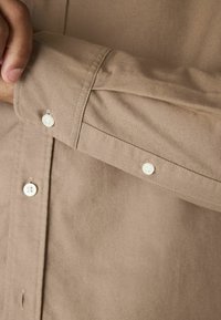 Lichtbruine blouse met lange mouwen, voorzien van een aangesloten manchette met witte knopen. Gladde stof met een subtiele textuur. Standaard kraagontwerp.