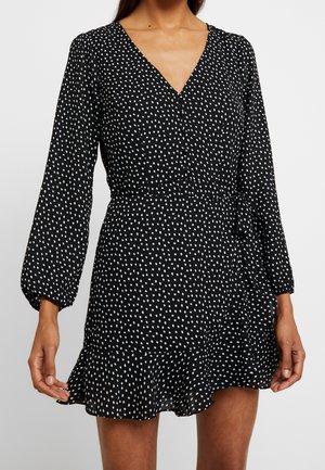 Black long-sleeve wrap dress with white polka dots, V-neckline, waist tie, and ruffled hem worn by a person shown from neck to mid-thigh.