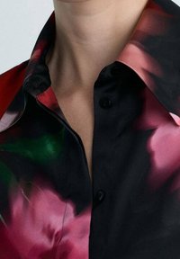 Chemise à col avec un fond noir orné de motifs floraux roses et verts vibrants, confectionnée en tissu lisse avec des boutons en détail.