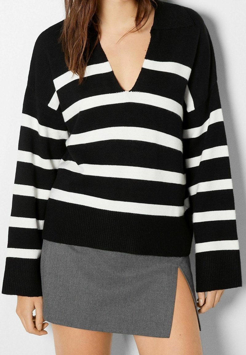Pull rayé noir et blanc avec un col en V profond et des manches longues, associé à une jupe grise avec une fente latérale. Tissu texturé.