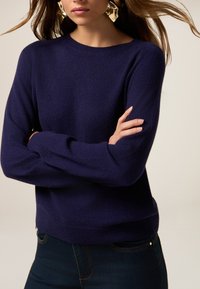 Granatowy sweter wykonany z miękkiego dzianinowego materiału, z klasycznym okrągłym dekoltem i długimi rękawami, w połączeniu z ciemnymi dżinsami.