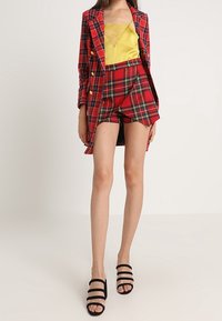 Rood-groen geruit colbert met gouden knopen, gele kanten cami, rode geruite shorts en zwarte sandalen met bandjes. Overal een gladde textuur.