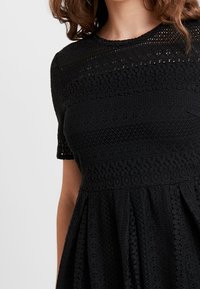 Schwarzes Spitzenkleid mit kurzen Ärmeln, das über komplexe Muster, einen strukturierten Oberkörper und einen ausgestellten Rock verfügt. Der Stoff ist leicht und mehrlagig.