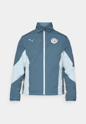 Windbreaker jacket in dark teal and light blue, featuring a zip front, high collar, and Manchester City logo on the chest.