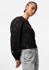 Melna caurspīdīga blūze ar ziedu mežģīņu rakstiem, pūstām piedurknēm un elastīgām aprocēm, kombinēta ar gaiši pelēnām augsta vidukļa džinsa biksēm.