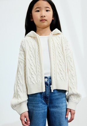 Weiße gestrickte Zip-Up-Strickjacke mit Kabelmuster, gerippten Bündchen und Kapuze, getragen über einem enganliegenden weißen Top und blauen Jeans.
