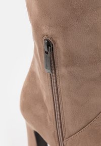Primer plano de una bota de ante marrón con una cremallera vertical y una tira de cuero con el nombre de la marca grabado.