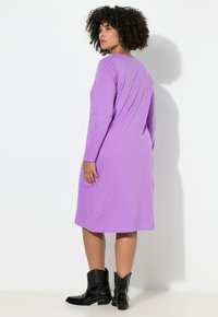 Fialové šaty s dlouhým rukávem a volnějším střihem, s hladkou texturou, délka ke kolenům, doplněné černými kotníkovými botami ve stylu kovbojů.