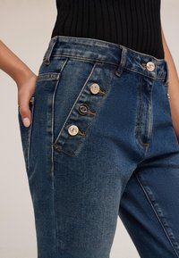 Detailní záběr osoby nosící modré džíny z denimu se šikmým řadovým uspořádáním tří stříbrných knoflíků a jednoho černého knoflíku poblíž přední kapsy.