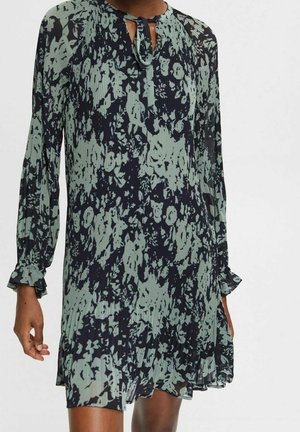 Robe longue à manches longues plissée avec motif floral abstrait noir et vert atténué, col noué, portée par une personne à la peau foncée.