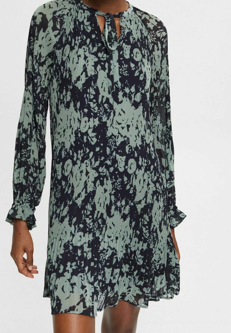 Robe longue à manches longues plissée avec motif floral abstrait noir et vert atténué, col noué, portée par une personne à la peau foncée.