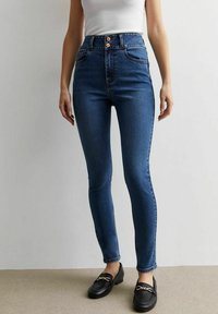 Jeans skinny de mezclilla azul de tiro alto con cierre de tres botones, cinco bolsillos y una textura suave. Llevados con mocasines negros.
