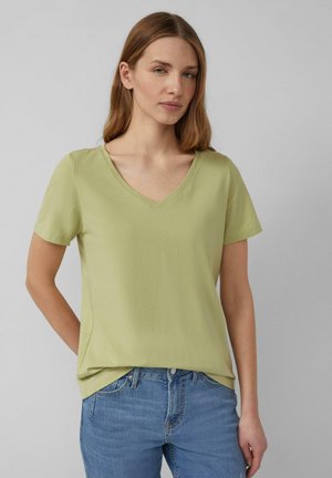 Vrouw met steil lichtbruin haar die een lichtgroen T-shirt met V-hals en korte mouwen en een blauwe spijkerbroek draagt, staand tegen een grijze achtergrond.