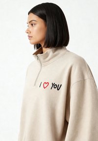 Sweatshirt beige à fermeture éclair avec texte "I ❤️ YOU" en noir et rouge sur le devant. Tissu doux et texturé avec un col montant.