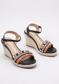 Sandali con zeppa nera e cinturini intrecciati nei colori arancione, bianco e nero. Le suole in stile espadrillas aggiungono texture e altezza. Chiusura con fibbia alla caviglia.