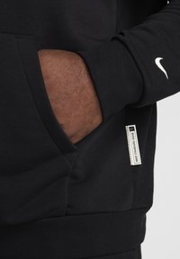 Felpa nera Nike con tasca a marsupio, polsini a coste e logo bianco sulla manica. Tessuto testurizzato con vestibilità casual.