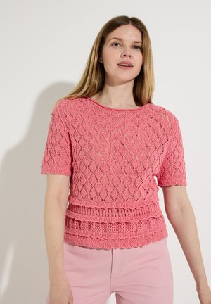 Femme portant un haut en maille rose à manches courtes avec des motifs texturés en losange et festonnés, associé à un pantalon rose clair, debout contre un mur blanc.