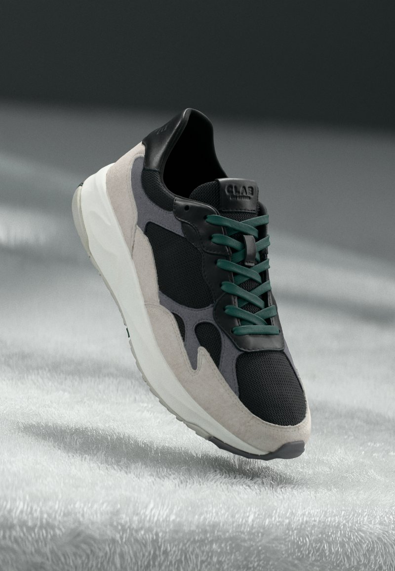 Black and gray sneaker with suede accents, mesh upper, green laces, and a white outsole; notable logo on the heel and streamlined design.