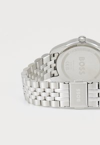 Montre-bracelet en acier inoxydable argenté avec un bracelet à maillons, un boîtier circulaire et un logo visible au dos. Résistante à l'eau jusqu'à 3 bars.