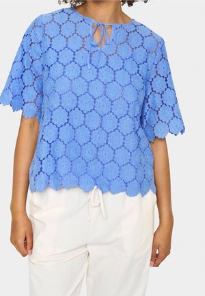 Personne portant un haut en dentelle bleu à manches courtes avec un motif floral et des bords festonnés, associé à un pantalon blanc à cordon.