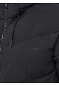 Gros plan d'une doudoune noire à coutures diagonales, avec fermeture éclair, cordon de serrage et logo discret embossé "Originals Studio" sur la poitrine.