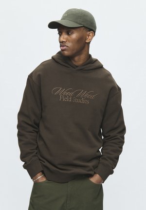 Jeune homme portant un sweat à capuche marron foncé "Wood Wood Field Studies" et une casquette vert olive, les mains dans les poches du pantalon vert, sur fond uni.