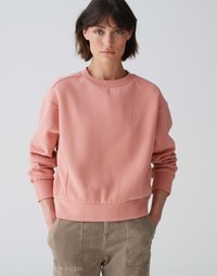 Felpa rosa con scollo rotondo, polsini a costine e un piccolo emblema a forma di cuore. Abbinata a pantaloni in velluto a coste di colore marrone chiaro. Tessuto morbido.