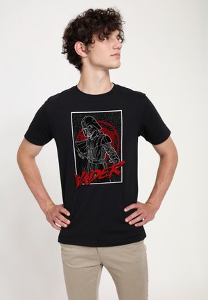 Junger Mann mit lockigem Haar schaut seitlich und trägt ein schwarzes T-Shirt mit einer weißen und roten Grafik von Darth Vader, der ein Lichtschwert hält, und dem Schriftzug "Vader".