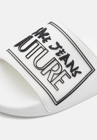 White slide sandal featuring a textured strap with raised black lettering reading "FUTURE" and "JEANS" in a rectangular outline. Soft sole.