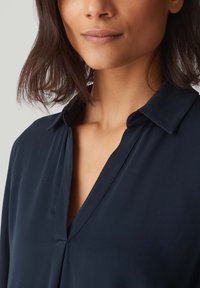 Blusa blu navy con scollatura a V e un tessuto morbido, caratterizzata da un colletto classico e una texture liscia. Adatta per essere indossata a strati o da sola.