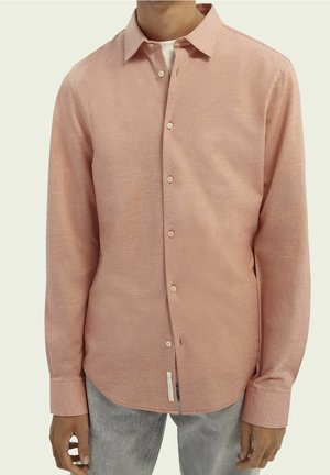 Mand iført langærmet, lyserød skjorte med knapper over en hvid undertrøje, kombineret med lysegrå jeans mod en ensfarvet baggrund.