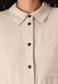 Camicia beige con colletto realizzata in tessuto texturizzato, dotata di bottoni neri e taschino sul petto, vista ravvicinata che mette in evidenza la pattina dei bottoni.
