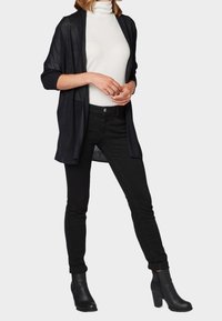 Svart öppensynt cardigan tillverkad av ett lätt, genomskinligt tyg, parat med en figurnära vit polotröja och svarta slim-fit jeans. Svarta ankelskor.