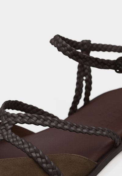 Σκούρα καφέ πλεγμένες δερμάτινες λουράκια που διασταυρώνονται πάνω από σανδάλι με επίπεδη σόλα, λείο πέλμα και κούμπωμα αστραγ άλου.