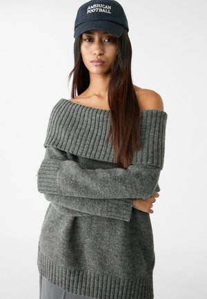 Grauer schulterfreier Pullover mit gerippter Textur und langen Ärmeln, ausgestattet mit einem weiten Ausschnitt, kombiniert mit einer dunkelblauen Mütze mit "American Football"-Logo.
