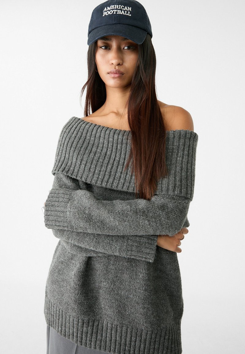 Grijze off-the-shoulder trui met ribbelstructuur en lange mouwen, met een wijde halslijn, gecombineerd met een donkerblauwe pet met het "American Football" logo.
