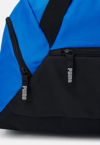 Sac de sport bleu et noir en nylon texturé, doté d'une poche zippée et d'une étiquette avec le logo argenté de Puma.