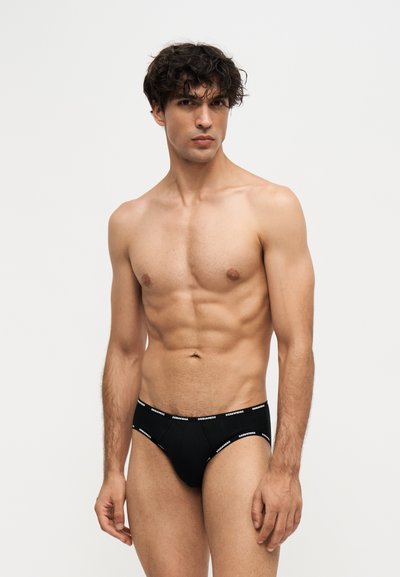 Jeune homme aux cheveux bouclés portant un slip noir avec un logo blanc sur la ceinture, debout devant un fond clair uni.