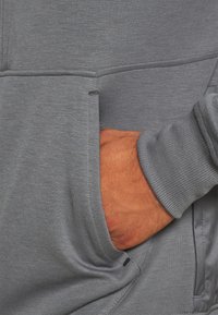 Main partiellement dans la poche avant d’un vêtement gris à manches longues avec coutures visibles et texture du tissu.