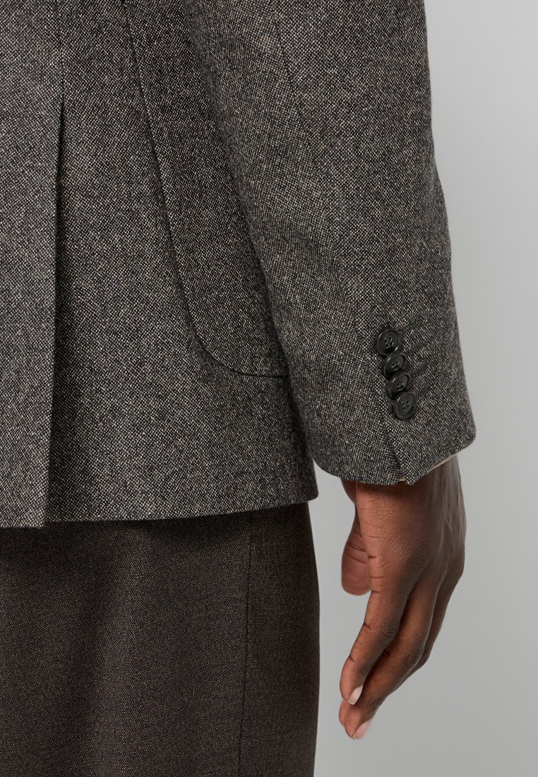 Nahaufnahme einer dunkelhäutigen Hand, die neben einem braunen Tweed-Blazerärmel mit vier schwarzen Knöpfen am Ärmel ruht.