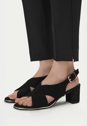 Pieds chaussés de sandales à talons blocs ouvertes en daim noir avec boucle dorée et breloque, associées à un pantalon noir ajusté à la cheville avec des fentes latérales.