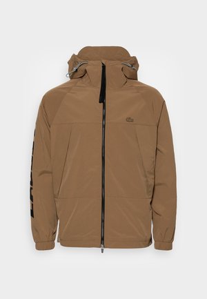 Brązowa kurtka z kapturem, zapinana na suwak z przodu, z elastycznymi mankietami, bocznymi kieszeniami oraz logo na lewej piersi i czarnym napisem na prawej rękawie.