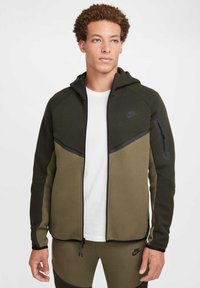Nike hooded jacket in olive green and dark green, featuring a zipper, side pockets, textured fabric, and black accent details.