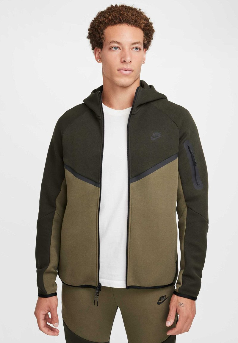 Giacca con cappuccio Nike in verde oliva e verde scuro, dotata di zip, tasche laterali, tessuto testurizzato e dettagli neri.