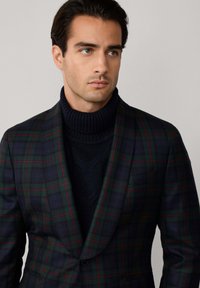 Uomo con capelli scuri che indossa un blazer a quadri verde e blu navy sopra un dolcevita nero, mentre guarda leggermente verso destra su uno sfondo semplice.