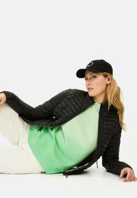 Black quilted jacket with long sleeves, paired with a green sweatshirt featuring a textured logo, and a black cap. Model is reclining.